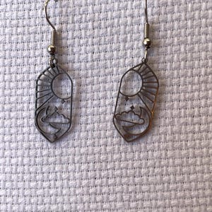 Peut inclure: Une paire de boucles d'oreilles de couleur bronze. Chaque boucle d'oreille présente un motif rectangulaire avec un soleil, une lune et des nuages. Les boucles d'oreilles ont une fermeture à crochet et sont présentées sur une surface blanche texturée.