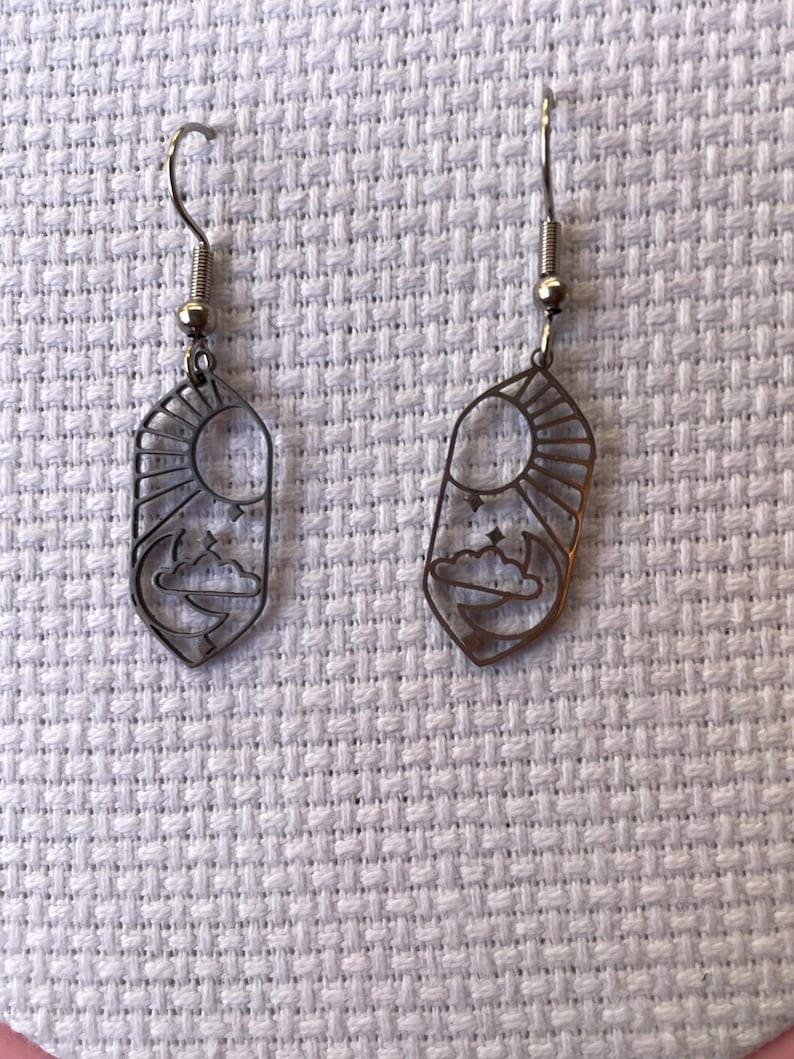 K&ouml;nnte beinhalten: Ein Paar bronzefarbene Ohrringe. Jeder Ohrring hat ein rechteckiges Design mit einem Sonnen-, Mond- und Wolkenmotiv. Die Ohrringe haben einen Hakenverschluss und werden auf einer wei&szlig;en, strukturierten Oberfl&auml;che pr&auml;sentiert.