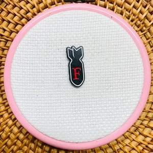 May include: A black enamel pin shaped like a bomb with a white outline and a red letter F in the centre, secured to a piece of white Aida fabric stretched within a circular pink embroidery hoop frame.