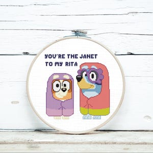 May include: A cross-stitch hoop with two cartoon dogs wearing hooded outfits. The text "YOU'RE THE JANET TO MY RITA" is stitched above the dogs. The dogs are in shades of purple, blue, orange, and green, against a white background.