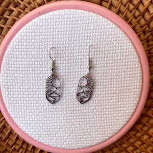 K&ouml;nnte beinhalten: Ein Paar silberfarbene H&auml;ngeohrringe. Jeder Ohrring hat ein ausgeschnittenes Design einer Sonne, Wolken und einer Bergkette. Die Ohrringe werden auf einem rosa Stickrahmen mit wei&szlig;em Stoff vor einem gewebten Hintergrund pr&auml;sentiert.