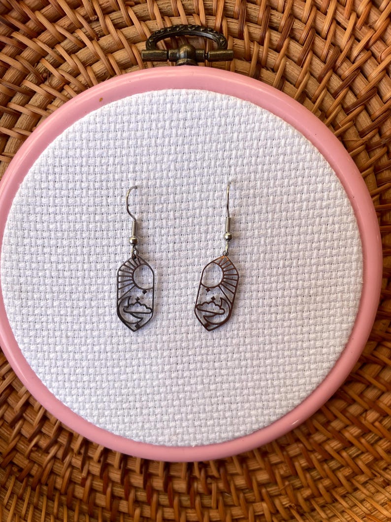 K&ouml;nnte beinhalten: Ein Paar silberfarbene H&auml;ngeohrringe. Jeder Ohrring hat ein ausgeschnittenes Design einer Sonne, Wolken und einer Bergkette. Die Ohrringe werden auf einem rosa Stickrahmen mit wei&szlig;em Stoff vor einem gewebten Hintergrund pr&auml;sentiert.