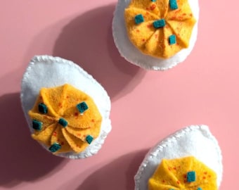 Pode incluir: Três ovos recheados de feltro sobre um fundo rosa. Cada ovo tem uma base branca e um recheio amarelo com detalhes em laranja e azul. Os ovos estão dispostos em um padrão triangular, projetando sombras.