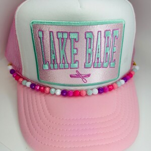 May include: Pink and white trucker hat with a green and purple embroidered patch that says "LAKE BABE" and a row of colorful beads around the brim.