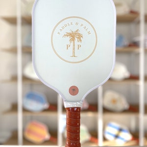 One side of a pickleball paddle, featuring the paddle and palm logo in yellow. The handle features chestnut brown grip tape.