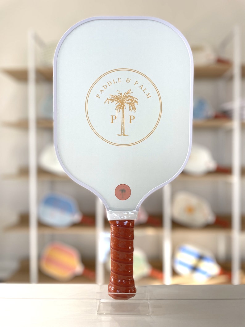 One side of a pickleball paddle, featuring the paddle and palm logo in yellow. The handle features chestnut brown grip tape.