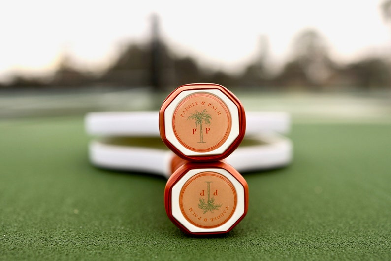 Two pickleball paddles laying on a pickleball court, showing the end cap of the handles which features the Paddle and Palm logo.