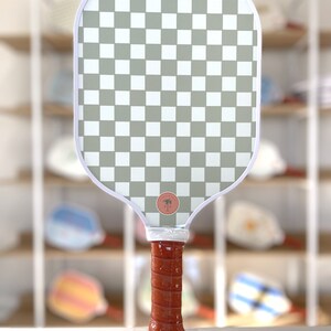 One side of a pickleball paddle, featuring a checkerboard pattern. The handle features chestnut brown grip tape.