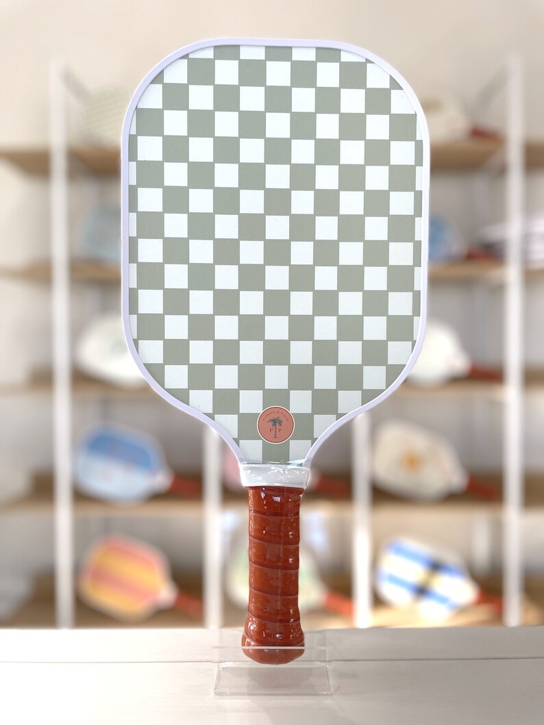 One side of a pickleball paddle, featuring a checkerboard pattern. The handle features chestnut brown grip tape.