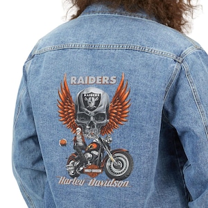 May include: A light blue denim jacket featuring a graphic on the back with a skull wearing a Raiders helmet, large orange wings, and a motorcycle. The text reads Raiders and Harley Davidson in a light-colored font against the blue fabric.