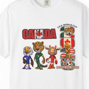 May include: White t-shirt featuring the word "CANADA" in red block letters, with the FIFA World Cup 2026 logo and mascot illustrations. The shirt includes the text "CANADA USA MEXICO".