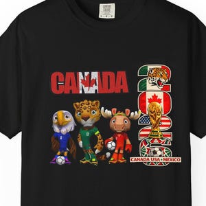 May include: Black t-shirt featuring the word "CANADA" in red, with the year "2026" and a graphic of the FIFA World Cup trophy. The shirt also displays cartoon animal mascots and the flags of Canada, Mexico, and the USA.
