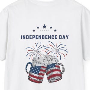 May include: A white t-shirt featuring a graphic of two beer mugs clinking together, each decorated with the American flag pattern. Above the mugs, the text reads HAPPY INDEPENDENCE DAY in blue, accompanied by red and blue firework illustrations.