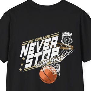 May include: A black short-sleeve t-shirt featuring a graphic of a basketball going through a hoop. The text reads NO FAILURE NEVER STOP JUMP SHOT with a small shield emblem containing the number 23 and the letters USA above the hoop.