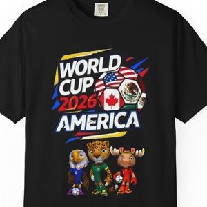 May include: Black t-shirt featuring the text "WORLD CUP 2026 AMERICA" with the flags of the United States, Canada, and Mexico. The shirt also displays cartoon mascots of an eagle, jaguar, and moose, each holding a soccer ball.
