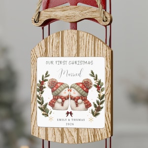 Op de afbeelding: Een houten slede-ornament met rode metalen glijders en een jute ophangkoord. Het midden heeft een wit paneel met een afbeelding van twee sneeuwpoppen met rode mutsen en sjaals, en de tekst "OUR FIRST CHRISTMAS Married", "EMILY & THOMAS", "2026".