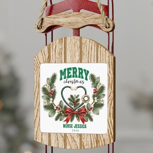 Op de afbeelding: Een decoratieve slede-ornament met een houtnerf-afwerking en rode metalen glijders. Het middenpaneel toont een krans met een stethoscoop in de vorm van een hart, een rode strik en de tekst Merry Christmas, Nurse Jessica, 2026.