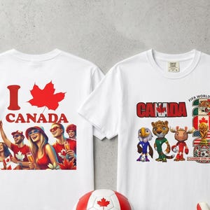 Op de afbeelding: Twee witte T-shirts met Canadese ontwerpen. E&eacute;n shirt toont "I Canada" met een esdoornblad en een feestelijke afbeelding. De andere toont "Canada" en FIFA World Cup graphics. Een rood-witte voetbal en keepershandschoenen zijn ook zichtbaar.