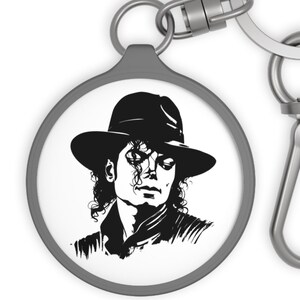May include: A round keychain featuring a black and white illustration of a person wearing a fedora hat. The keychain has a silver-colored ring and clasp for attaching to keys or bags. The image is a portrait.