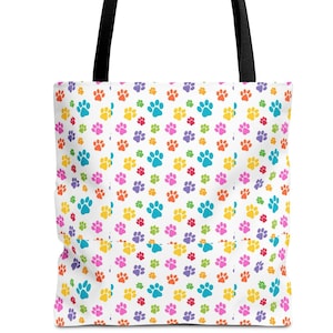 May include: White tote bag with a repeating pattern of colorful paw prints in blue, yellow, pink, purple, and green. The bag has black handles.
