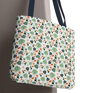 May include: A white tote bag with a repeating pattern of toucans, green leaves, and flowers. The bag has navy blue handles and is hanging against a neutral-colored wall. The design features orange, black, and pink accents.