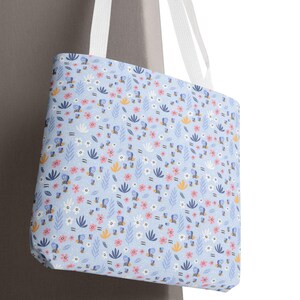 May include: A light blue tote bag with a floral pattern. The pattern includes pink, white, and yellow flowers, along with blue and green leaves. The bag has white handles.
