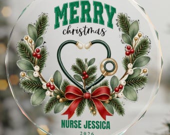 Op de afbeelding: Een ronde, doorzichtige glazen kerstornament met een groen en rood kransontwerp en een stethoscoop in de vorm van een hart. De tekst luidt MERRY christmas, NURSE JESSICA en 2026. Het hangt aan een dun rood lint tegen een wazige boom.