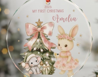 Op de afbeelding: Een ronde, doorzichtige glazen kerstornament hangend aan een rood lint. Het ontwerp toont een groene kerstboom met een roze strik, een kleine olifant en een konijn in een bloemenjurk. De tekst luidt 2026 MY FIRST CHRISTMAS en Amelia.