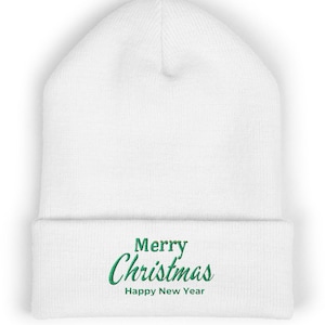 Op de afbeelding: Een witte gebreide muts met een omgeslagen boord en de geborduurde groene tekst Merry Christmas Happy New Year in het midden aan de voorzijde. De muts is gemaakt van een zachte stof met een zichtbare ribbelstructuur over het geheel.