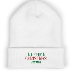 Op de afbeelding: Een witte muts met omslag voorzien van geborduurde tekst Merry Christmas Jessica in rode en groene letters, geflankeerd door twee kleine groene kerstboomiconen op de voorzijde van de omgeslagen rand.