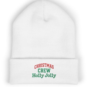 Op de afbeelding: Een witte muts met omslag en geborduurde tekst op de voorzijde. Het woord CHRISTMAS is in het rood gestikt, terwijl de woorden CREW en Holly Jolly in het groen gecentreerd zijn op de omgeslagen rand van de gebreide muts.