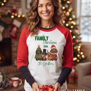 May include: A white long-sleeve shirt with red raglan sleeves and black cuffs featuring a graphic of four dachshunds in holiday attire, the text "FAMILY Christmas 2026" and "The Williams" printed in green, and a small gift box held in front.