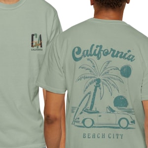 May include: A sage green short-sleeve t-shirt featuring a small CA California logo on the front chest. The back displays a graphic of a convertible car, a surfboard, a palm tree, and two suns with the text California Beach City in dark green.