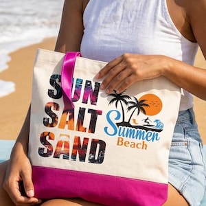May include: A cream-colored canvas tote bag with a vibrant pink bottom panel. The front features the text SUN SALT SAND in a sunset-themed font, alongside a graphic of palm trees, a sun, and the blue text Summer Beach with a small surfer icon.