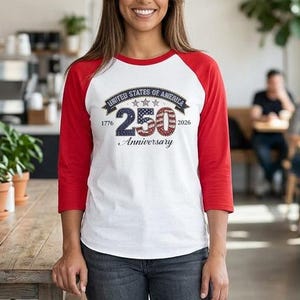 May include: A white and red baseball tee with the text "UNITED STATES OF AMERICA 250th Anniversary 1776-2026". The shirt is worn by a person in a cafe setting. The shirt has red sleeves and a white body.