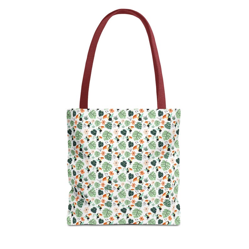 May include: White tote bag with a tropical pattern of toucans, flowers, and leaves. The bag has a burgundy handle. The pattern includes orange, green, and pink elements. The bag is designed for carrying items.