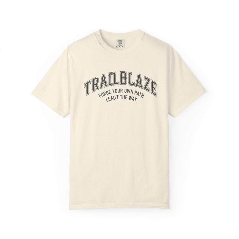 May include: Cream-colored t-shirt with the word "TRAILBLAZE" arched across the chest in a dark gray, collegiate-style font. Below, the text reads "FORGE YOUR OWN PATH LEAD THE WAY" in a smaller font. The shirt has a crew neck and short sleeves.