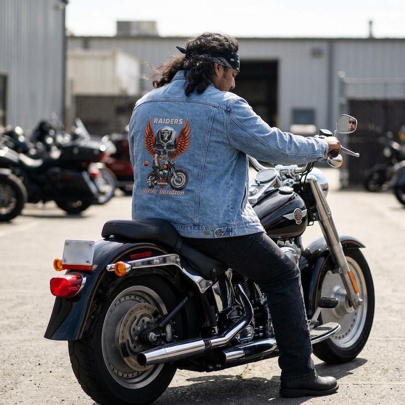 May include: A light blue denim jacket featuring a graphic on the back with a skull, orange wings, and the text Raiders and Harley Davidson. The jacket is worn over dark pants while sitting on a black motorcycle with chrome exhaust pipes.