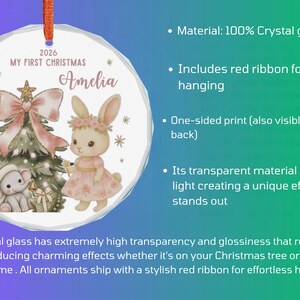 May include: A round crystal glass ornament featuring a printed illustration of a Christmas tree, a small elephant, and a rabbit wearing a floral crown. The text reads 2026 MY FIRST CHRISTMAS and the name Amelia. It includes a red ribbon for hanging.