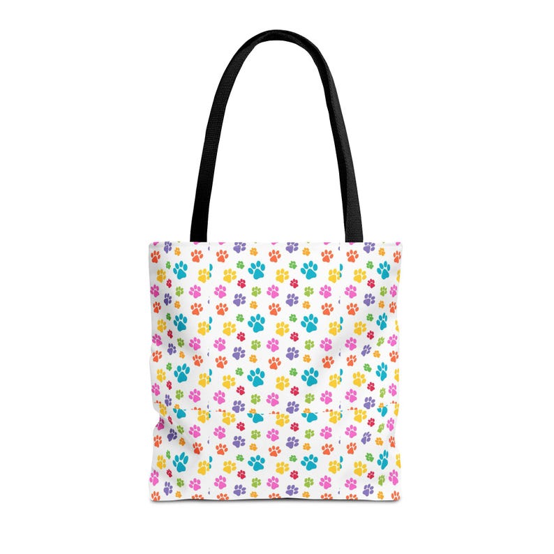 May include: White tote bag with a repeating pattern of colorful paw prints in blue, yellow, pink, purple, and green. The bag has black handles.