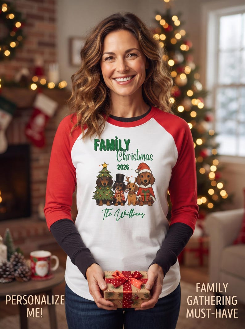 May include: A white long-sleeve shirt with red raglan sleeves and black cuffs featuring a graphic of four dachshunds in holiday attire, the text "FAMILY Christmas 2026" and "The Williams" printed in green, and a small gift box held in front.