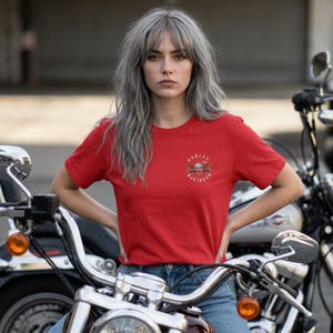K&ouml;nnte beinhalten: Ein leuchtend rotes Kurzarm-T-Shirt mit einem kleinen Harley-Davidson-Logo auf der linken Brust. Das Logo zeigt eine Grafik eines Totenkopfes mit Fl&uuml;geln und den Schriftzug Harley-Davidson in kreisf&ouml;rmiger Anordnung. Mit Rundhalsausschnitt.