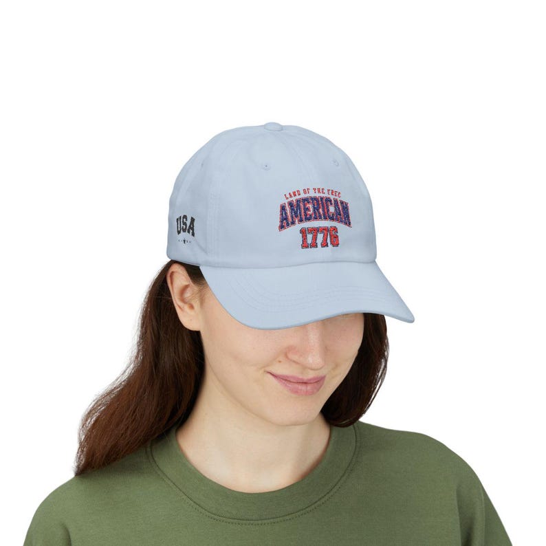 K&ouml;nnte beinhalten: Eine hellblaue Baseballkappe mit dem Text LAND OF THE FREE AMERICAN 1776 in Rot, Wei&szlig; und Blau auf der Vorderseite. An der Seite der Kappe befindet sich der Text USA mit einem kleinen Sternsymbol. Die Kappe hat einen gebogenen Schirm und eine strukturierte Krone.