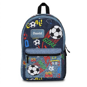 May include: A denim-textured backpack featuring a soccer-themed print with black and white soccer balls, red and green text reading GOAL, and the name David printed on a rectangular patch. The bag includes a front zippered pocket and black trim.