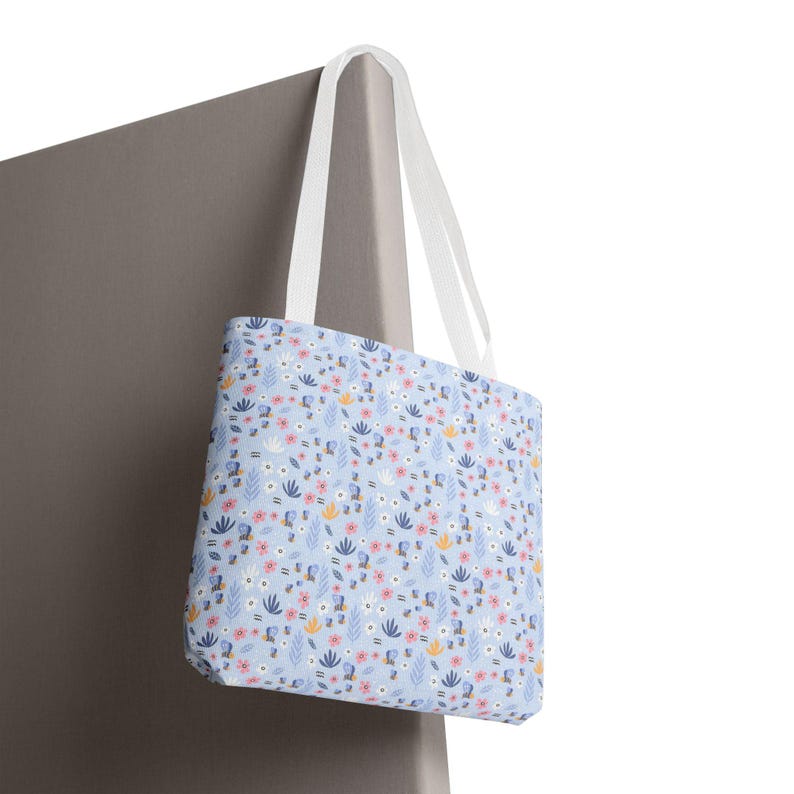 May include: A light blue tote bag with a floral pattern. The pattern includes pink, white, and yellow flowers, along with blue and green leaves. The bag has white handles.