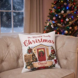 May include: A white square throw pillow featuring a festive holiday illustration of a house entrance with a wreath, Christmas trees, and the text The BROWN Family Christmas, David, and 2027 printed in red and brown script and serif fonts.