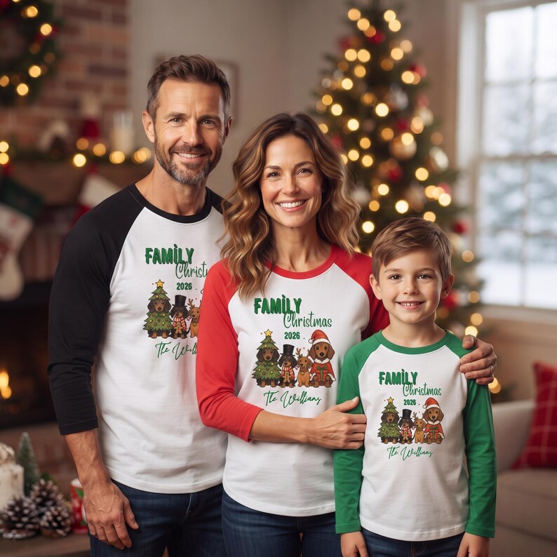 Op de afbeelding: Drie raglan-shirts met een witte romp en mouwen in zwart, rood en groen. Elk shirt heeft een afbeelding van vier teckels in feestelijke kleding, de tekst "FAMILY CHRISTMAS 2026" en de handgeschreven naam "The Williams".