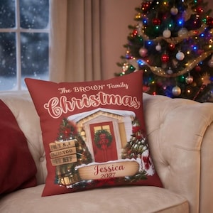 May include: A square red throw pillow featuring a festive illustration of a house entrance with a wreath on a red door, flanked by two Christmas trees. The text reads The BROWN Family Christmas, with a wooden sign displaying the name Jessica 2027.