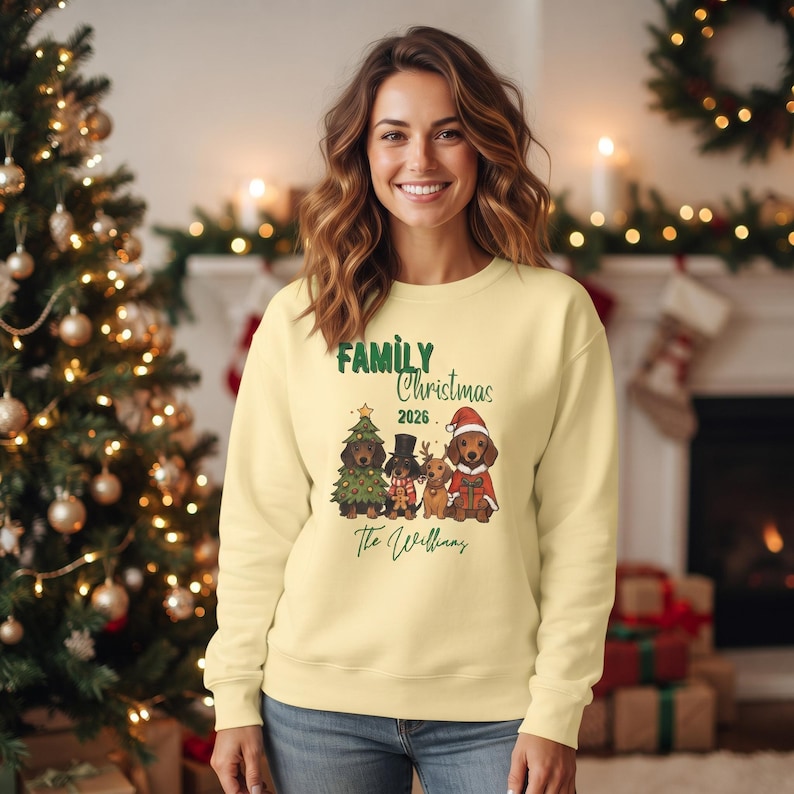 May include: A pale yellow crewneck sweatshirt featuring a graphic of four dachshunds in holiday attire. The text reads Family Christmas 2026 and The Williams. One dog wears a tree costume, one a top hat, one reindeer antlers, and one a Santa suit.