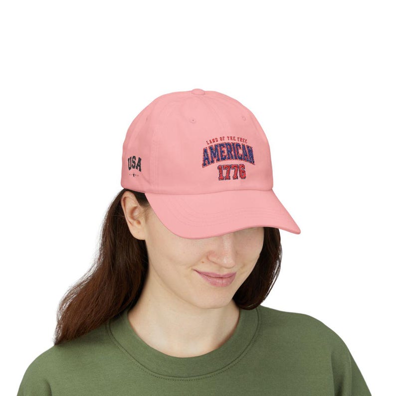 K&ouml;nnte beinhalten: Eine hellrosa Baseballkappe mit dem Text LAND OF THE FREE AMERICAN 1776 in einer blau-roten Schriftart auf der Vorderseite. An der Seite der Kappe befindet sich der Text USA in schwarzer Schrift. Die Kappe hat einen gebogenen Schirm und eine feste Form.